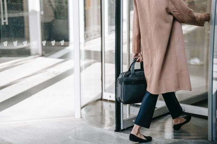 Woman wearing a pale pink coat, tailored jeans and ballet flats, and holding a black handbag, enters a revolving door. We are only able to see up to her shoulders.
